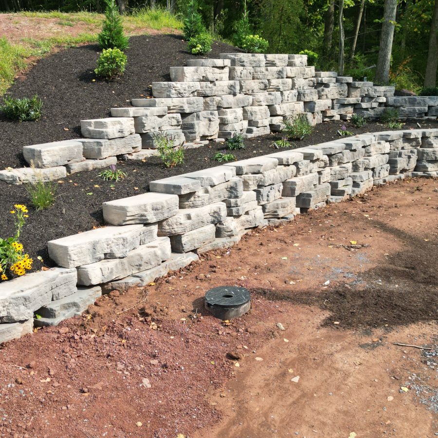 Left angle hardscaped retaining wall at the edge of property with landscaped trees and shrubs