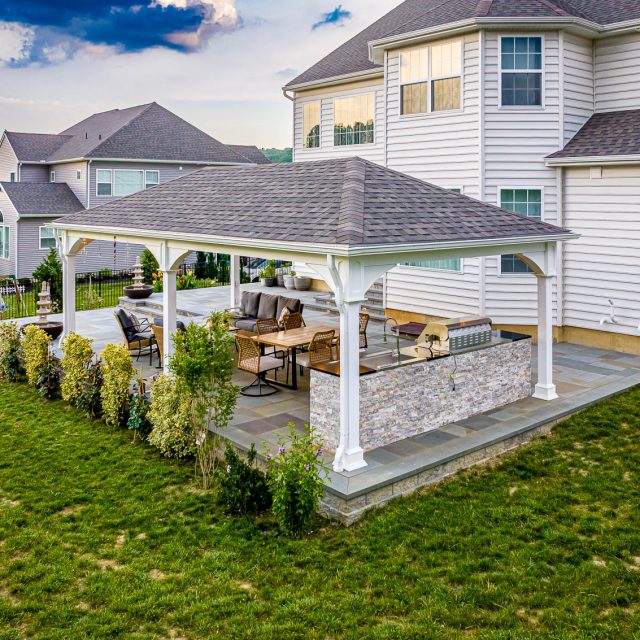 Backyard paver patio with white pavilion, outdoor dining area, seating space, and stone outdoor kitchen beside a house.