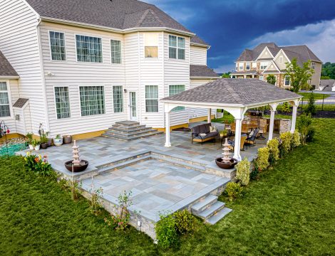 Backyard paver patio with tiered layout, stone steps, and white pavilion seating area behind a two-story home.
