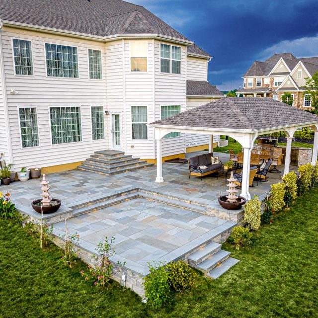 Backyard paver patio with tiered layout, stone steps, and white pavilion seating area behind a two-story home.