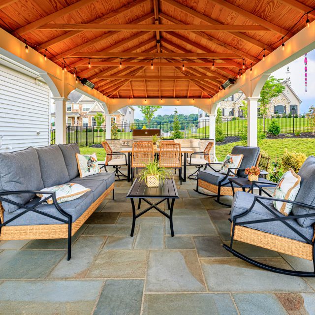 Outdoor seating area beneath a wood ceiling pavilion with patio furniture, string lights, and paver patio flooring.
