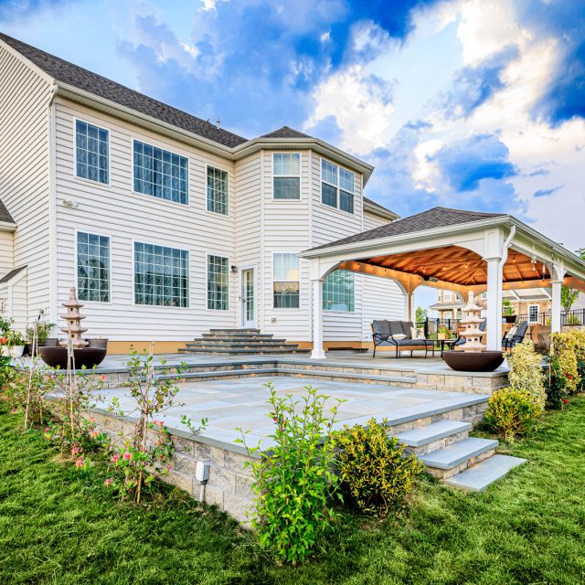 Backyard paver patio with tiered stone steps, white pavilion seating area, and landscaping beside a two-story home.