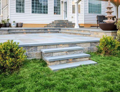 Backyard paver patio with tiered stone steps, built-in lighting, and landscaping beside a house.