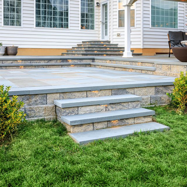 Backyard paver patio with tiered stone steps, built-in lighting, and landscaping beside a house.
