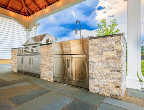 Outdoor kitchen with stainless steel grill, sink, and stone veneer island beneath a white pavilion on a paver patio.