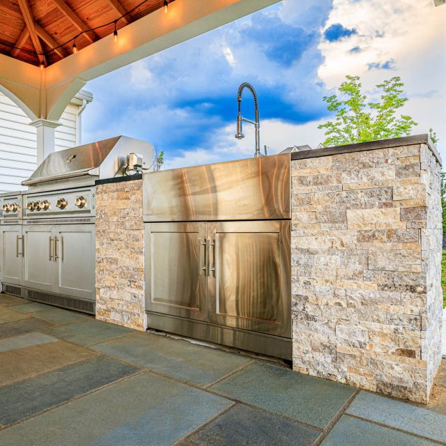 Outdoor kitchen with stainless steel grill, sink, and stone veneer island beneath a white pavilion on a paver patio.