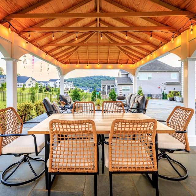 Outdoor dining area beneath a wood pavilion with string lights, wicker chairs, and large paver patio.