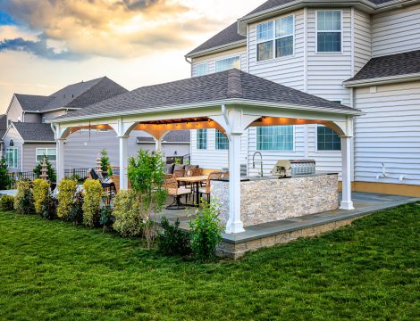 Backyard pavilion with outdoor kitchen, dining area, and large paver patio beside a two-story home.