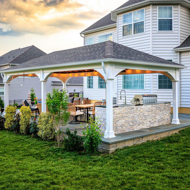 Backyard pavilion with outdoor kitchen, dining area, and large paver patio beside a two-story home.