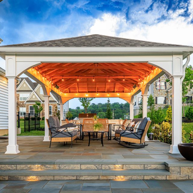 White backyard pavilion with string lights, outdoor dining set, and paver patio with illuminated steps.