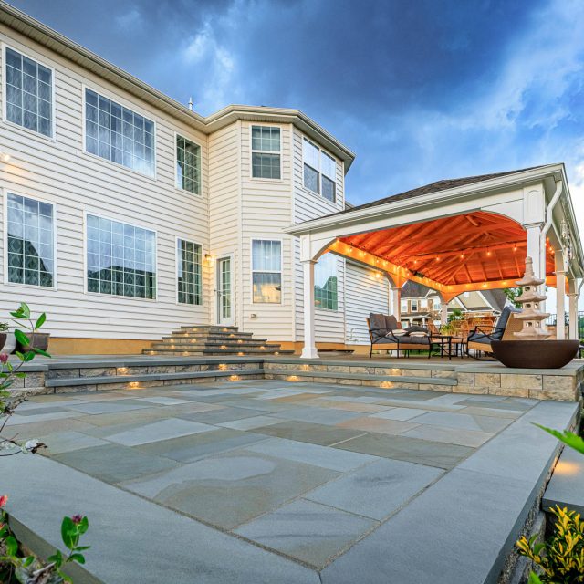 Large backyard paver patio with illuminated stone steps and white pavilion seating area beside a two-story home.
