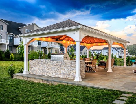 Backyard pavilion with outdoor kitchen, dining area, and large paver patio beside residential homes.