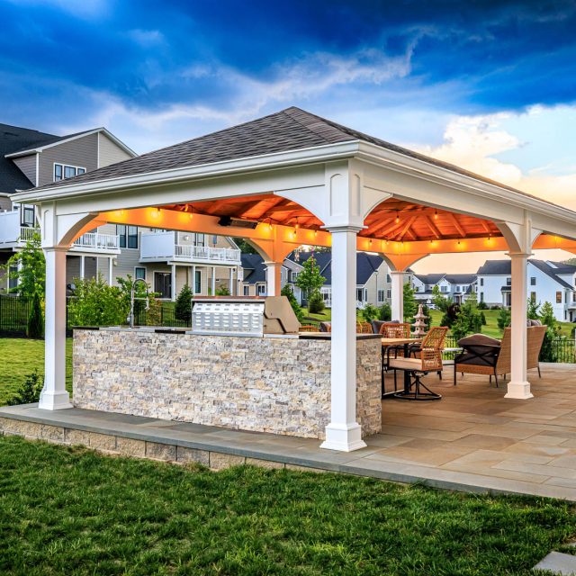 Backyard pavilion with outdoor kitchen, dining area, and large paver patio beside residential homes.