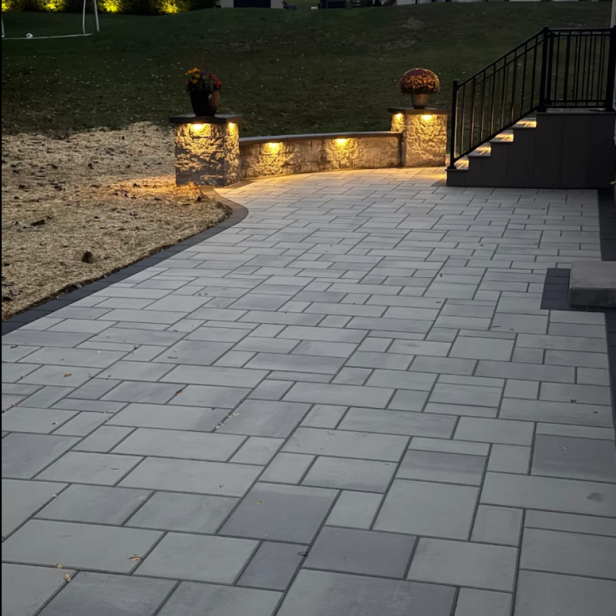 Gray paver patio with curved stone wall, pillar lights, and stair lighting next to raised deck at dusk.