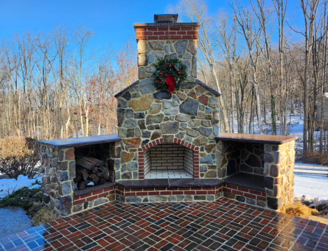 Stone outdoor fireplace with brick firebox, wood storage niches, chimney, and brick patio surrounded by trees.